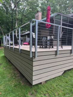 Corner of a raised composite deck with gray aluminum posts and stainless cable railing on a lawn.