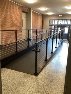 Black posts with cable infill and grab rails line an interior ramp in a renovated brick building.