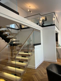 Light oak floating treads with glowing under-tread LEDs climb beside frameless glass with a wood cap rail to a glass-railed loft.