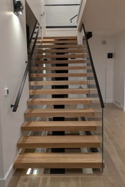 Straight run of white oak floating treads on a mono stringer with frameless glass on one side and a black wall-mounted handrail on the other.