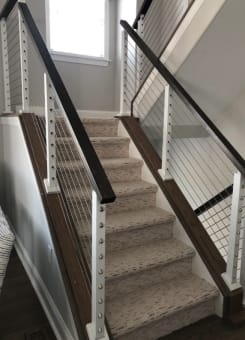 Black top rail and white posts with stainless cable infill mounted on knee walls flanking a carpeted stair.