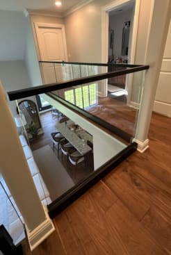 Upper hallway with frameless glass guard topped with a black cap rail overlooking a dining room.