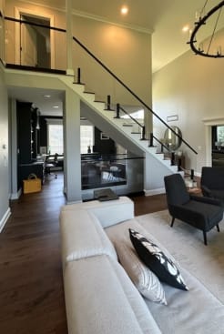 Open great room with a white-riser stair lined by frameless glass on black talons and a black handrail, plus a glass loft railing above.