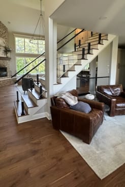 Wood stair with white risers and frameless glass on black talons rising beside a leather-furnished family room.