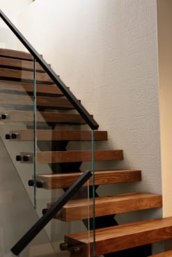Walnut floating treads rise along a textured white wall with frameless glass and a black handrail.