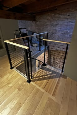 Black horizontal rod railing with a maple handrail surrounds a stair opening in a brick-walled loft.