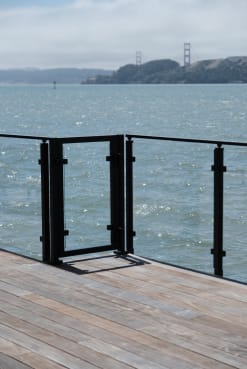 Black post-to-post glass railing and matching gate on a wood dock deck overlooking the bay and a distant bridge.