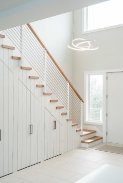 A white board-and-batten stair with oak treads and slim white cable railing beneath a ring pendant light.