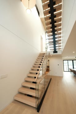 Light oak treads climb a black mono stringer through a narrow triple-height space with glass railing.