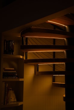 Curved wood spiral treads glow with LED strips in a dark room beside built-in bookshelves.