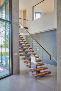 Rich wood floating treads on a mono stringer rise past a board-formed concrete column with frameless glass and a wood handrail.
