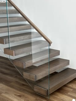 Thick gray-brown floating treads project from a white wall with frameless glass and a wood handrail.