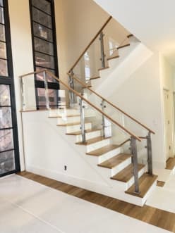 Brushed stainless posts hold glass panels with an oak handrail along a white-and-oak switchback stair beside tall black-framed windows.