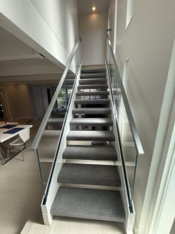Frameless glass guards with slim metal handrails line both sides of a straight gray-carpeted stair.