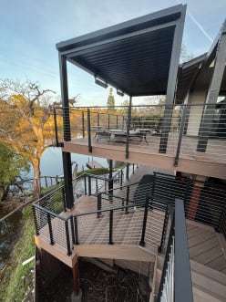 Black-post cable railing wraps a two-level wood deck and stair overlooking a lake at sunset.