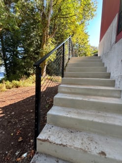 Black rod railing with slim side-mounted posts climbs a concrete stair along a wooded hillside.