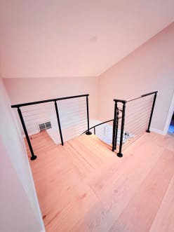 Black posts with cable infill guard a stair opening on light hardwood floors.