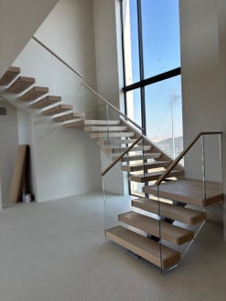 Light oak floating treads with frameless glass and wood handrail rise beside a tall window wall.