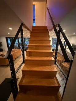 Black cable railings line both sides of a straight wood stair leading down into a modern living room.