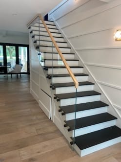 Frameless glass on black standoff pins with an oak handrail beside black treads and white risers.