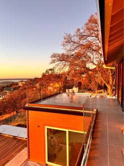 Frameless glass railing runs along a rooftop terrace overlooking oak trees and a valley at sunset.