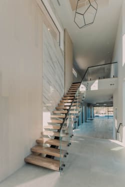 Oak treads on a black mono stringer with standoff-pin glass rise along a marble wall in a long bright hall.