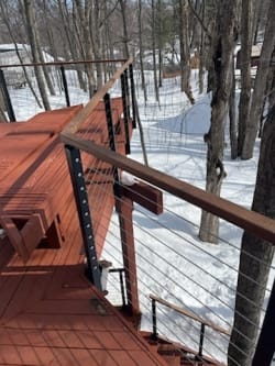 Black side-mount posts with cable infill and a wood cap rail on a red-stained deck in snowy woods.