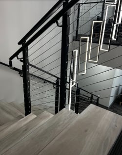 Black cable railing with wall handrail along a switchback stair under hanging rectangular pendant lights.