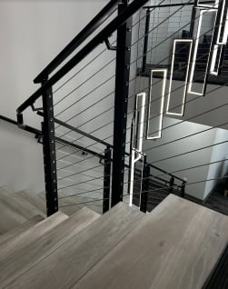 Black cable railing with wall handrail along a switchback stair under hanging rectangular pendant lights.