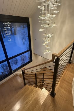 Oak stair landing with onyx black rod railing below a cascading glass chandelier and snowy windows
