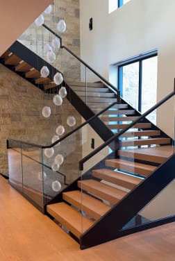 Black FLIGHT switchback with oak treads and glass railing beside a stone wall under cascading glass pendants.