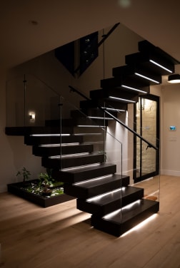Dark-stained floating treads with under-tread LEDs and frameless glass railing rise above an indoor planter bed.