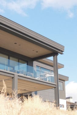 Frameless glass railing lines a covered second-story balcony seen from tall hillside grasses.