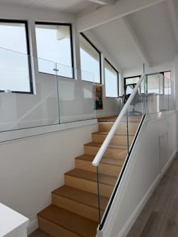 Frameless glass with a slim white handrail follows an oak stair up to a glass-guarded loft below angled clerestory windows.