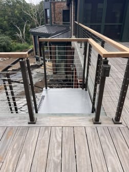 Black cable railing with wood cap frames a stair opening on a weathered wood deck.