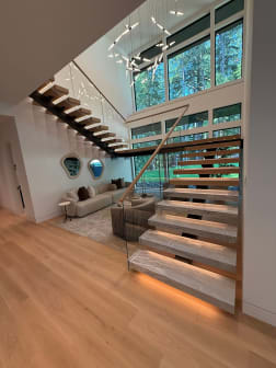 Floating stone-look treads with LED lighting and glass railing rise along a wall of windows in a vaulted living room.