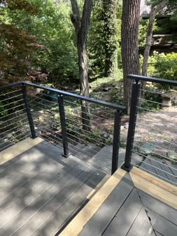 Black cable railing frames deck steps leading down into a shaded wooded garden.