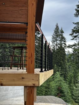 Black cable railing runs along a raised cedar deck supported by timber posts in a pine forest.