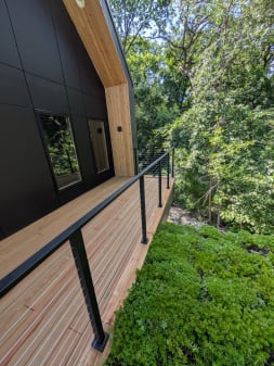 Black cable railing runs along a cedar deck of a black modern home beside lush trees.