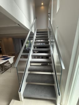 Frameless glass guards with slim metal handrails line both sides of a straight gray-carpeted stair.