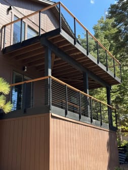 Two stacked decks with black posts, wood caps and cable infill project from a brown cabin among tall pines.