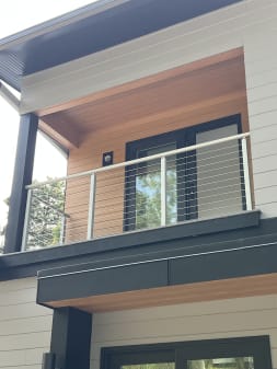 A polished stainless cable railing with round handrail guards a recessed balcony with wood soffit on a white modern home.