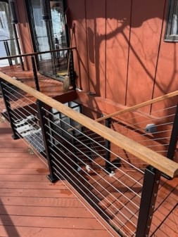 An ipe wood handrail tops black cable railing on a red-stained deck stair.