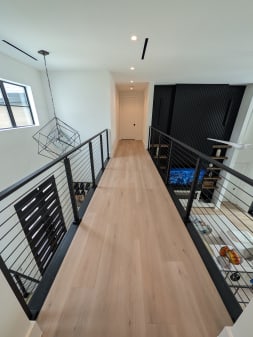 Upper floor oak bridge walkway lined on both sides by black cable railing above an open living area.