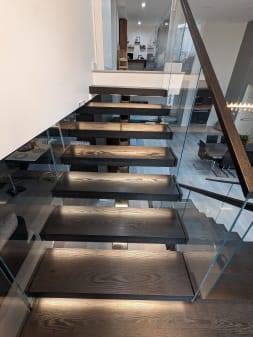 Dark-stained oak floating treads with LED under-lighting flanked by frameless glass and a wood handrail.