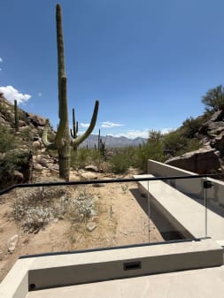 Frameless glass with a black top rail guards a concrete terrace among saguaro cacti and rocky hills.