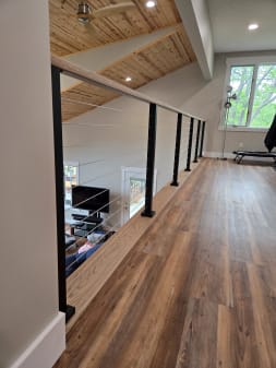 Black-post cable railing with a wood handrail runs along a wood-floored loft overlooking the room below.