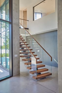 Floating wood treads on a center stringer with frameless glass and a wood handrail beside a concrete column and tall windows.