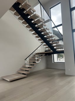 Light oak floating treads on black mono stringers with frameless glass railing in a bright double-height room.
