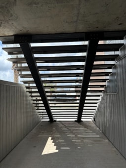 Looking up beneath a wide exterior steel stair with twin black stringers and open treads casting striped shadows.